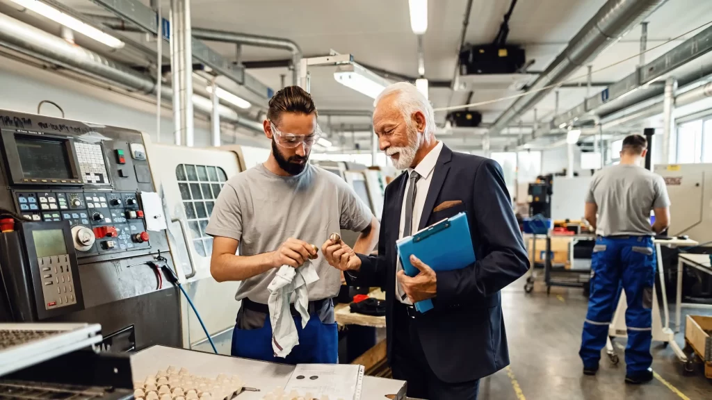 Senior inspector inspecting a part of machine in factory with young worker